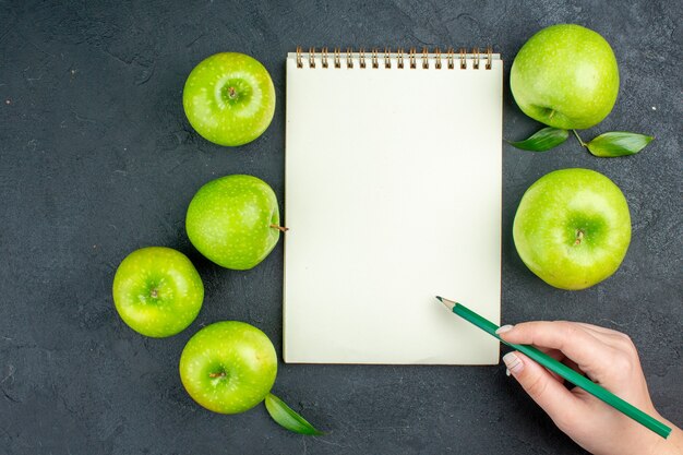 Vista superior cuaderno verde manzanas lápiz verde en mano femenina sobre superficie oscura