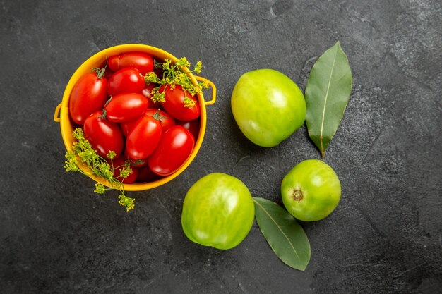 Vista superior del cubo amarillo lleno de tomates cherry y flores de eneldo, hojas de laurel y tomates verdes sobre un suelo oscuro con espacio de copia