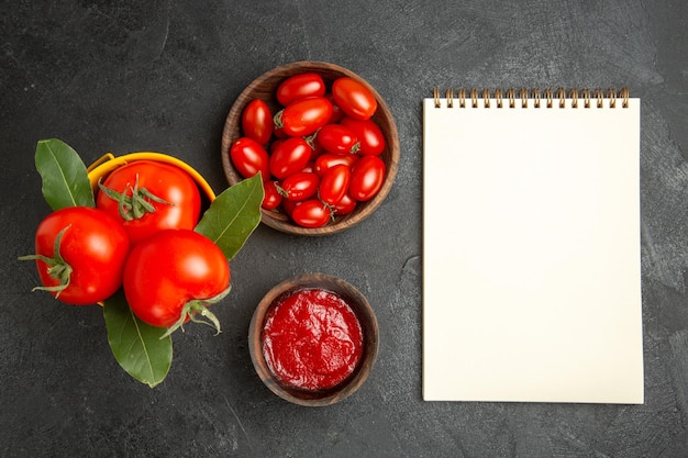 Vista superior del cubo amarillo con tomates y hojas de laurel, cuencos con tomates cherry y salsa de tomate y un cuaderno sobre suelo oscuro