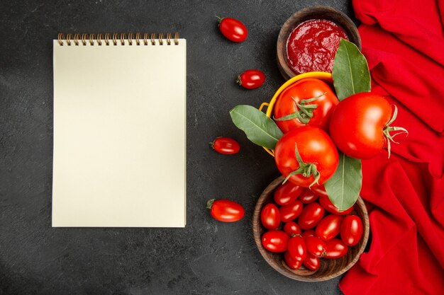 Vista superior de un cubo con tomates y hojas de laurel, cuencos con tomates cherry y ketchup rojo toalla un cuaderno sobre suelo oscuro