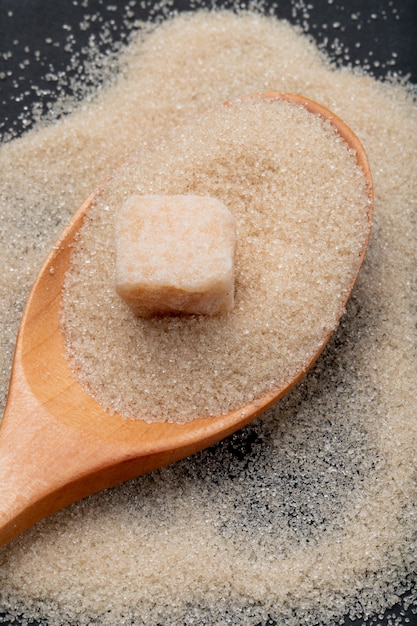 Vista superior de una cuchara de madera con azúcar moreno granulado y terrón de azúcar sobre fondo negro
