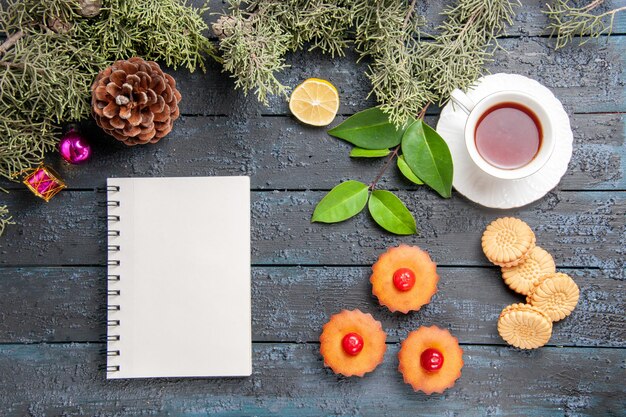 Vista superior de cupcakes de cereza ramas de abeto hojas de rodaja de limón, una taza de té, galletas y un cuaderno en la mesa de madera oscura.