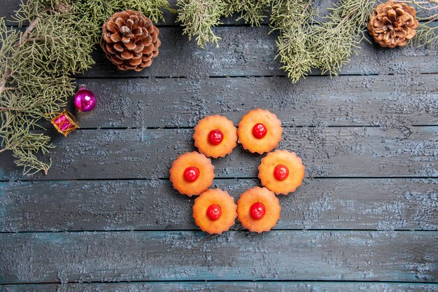 Vista superior de cupcakes de cereza redondeados ramas de abeto juguetes de navidad y piñas en suelo de madera oscura con espacio de copia