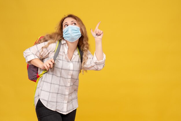 Vista superior de la curiosa chica viajera con su máscara y mochila apuntando hacia arriba en amarillo