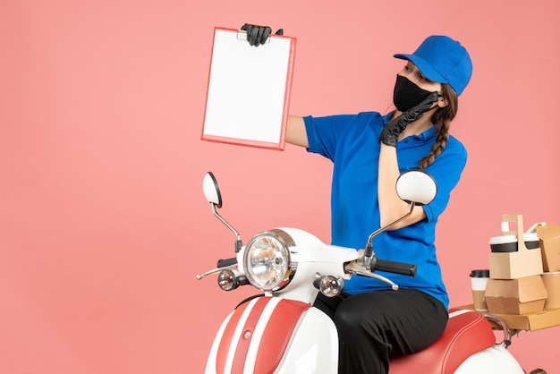 Vista superior de la curiosa mujer de mensajería con máscara médica y guantes sentado en scooter sosteniendo hojas de papel vacías entregando pedidos sobre fondo melocotón pastel