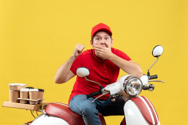 Vista superior del curioso adulto joven con blusa roja y sombrero entregando orden apuntando hacia atrás sobre fondo amarillo