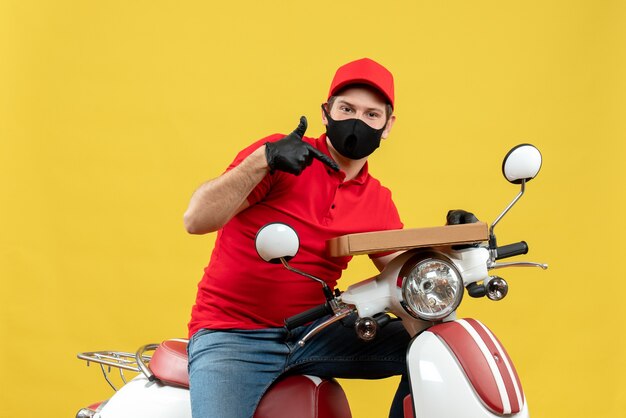 Vista superior del curioso mensajero hombre vestido con blusa roja y guantes de sombrero en máscara médica sentado en scooter mostrando orden