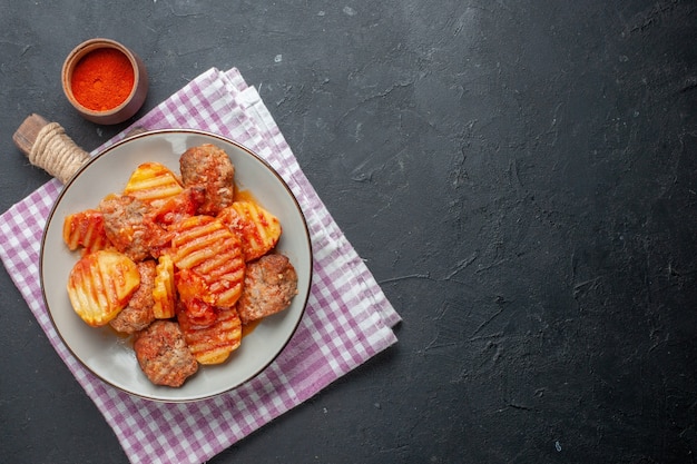 Vista superior de una deliciosa cena con papa y tomate de carne en una toalla despojada de color púrpura y pimiento rojo en el lado derecho sobre negro