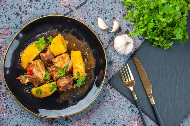 Vista superior de la deliciosa cena con patatas de carne servidas con verde en un plato negro y cubiertos en la tabla de cortar ajo sobre fondo de colores mezcla