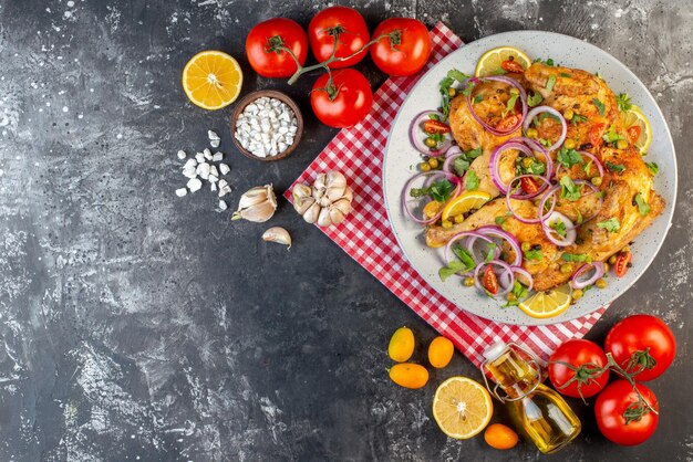 Vista superior de la deliciosa cena plato de pollo frito con varias especias y alimentos tomates con tallos ajos botella de aceite caída limón en el lado izquierdo sobre fondo de color oscuro