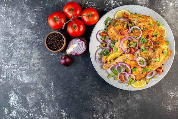 Vista superior de una deliciosa cena plato de pollo frito con varias especias y alimentos tomates con tallos pimientos cebollas en el lado izquierdo sobre fondo de color oscuro