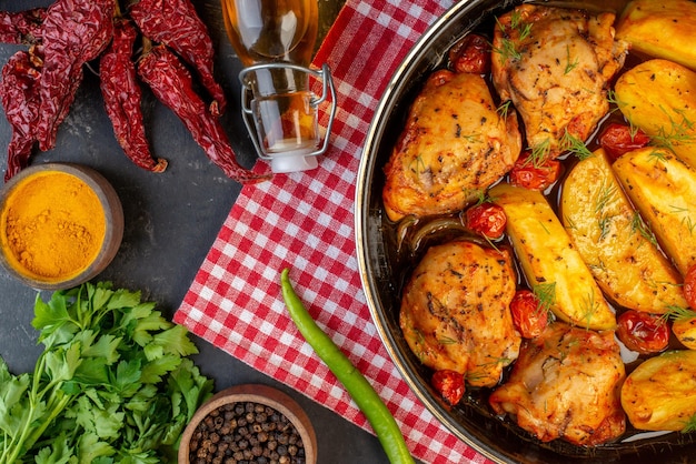 Vista superior de una deliciosa cena con pollos, papas, verduras sobre una toalla roja doblada, pimientos secos, una botella de aceite caída, un paquete verde de jengibre amarillo en el lado derecho sobre un fondo de color oscuro