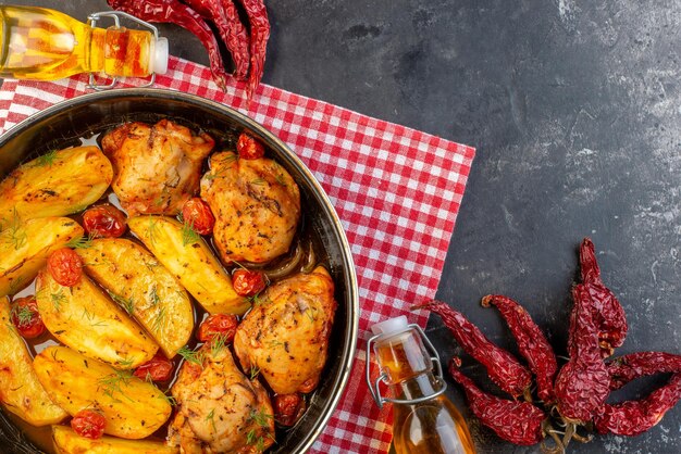 Vista superior de una deliciosa cena con pollos, patatas, verduras en una cacerola sobre una toalla roja doblada, pimientos secos, botella de aceite caída en el lado derecho sobre fondo de color oscuro