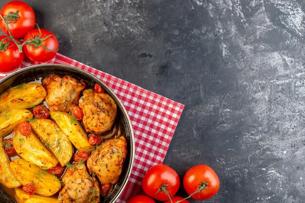 Vista superior de una deliciosa cena con pollos, patatas, verduras en una cacerola sobre una toalla roja doblada, tomates frescos sobre fondo de color oscuro