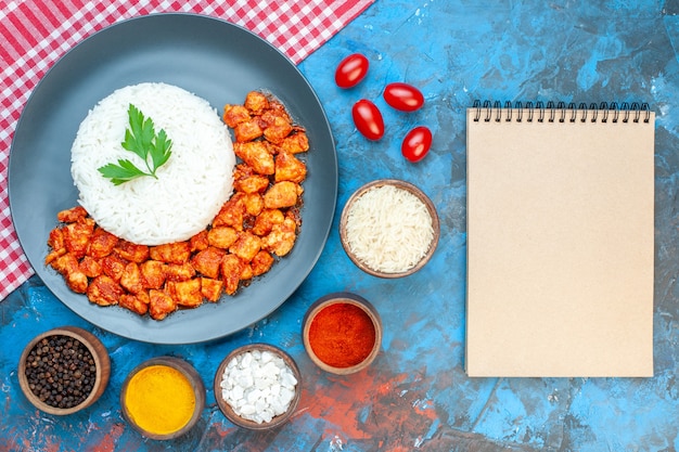 Vista superior de la deliciosa comida de arroz con pollo verde y tomate en una toalla despojada roja tomates de diferentes especias junto al cuaderno de espiral en la mesa azul