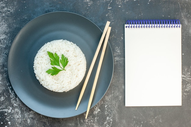 Vista superior de la deliciosa comida de arroz servida con palillos verdes y en un plato negro junto al cuaderno de espiral sobre fondo oscuro