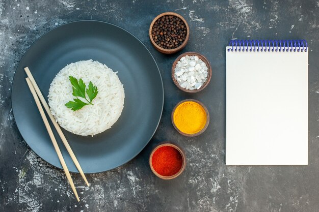 Vista superior de la deliciosa comida de arroz servida con palillos verdes y en un plato negro junto a diferentes especias junto al cuaderno de espiral en el lado derecho sobre fondo oscuro