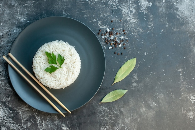 Vista superior de la deliciosa comida de arroz servida con palillos verdes y en un plato negro pimientos en el lado derecho sobre fondo oscuro