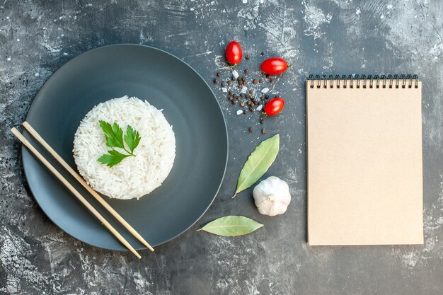 Vista superior de la deliciosa comida de arroz servida con palillos verdes y en un plato negro pimientos tomates al ajillo cuaderno de espiral sobre fondo oscuro