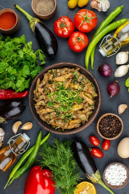 Vista superior de la deliciosa comida de berenjena, verduras frescas, especias, botella de aceite caída, paquete verde sobre fondo de color oscuro