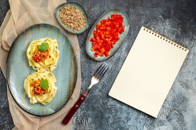 Vista superior de una deliciosa comida de pasta en un plato azul servido con tomate y carne para la cena en una toalla de color bronceado con sus ingredientes junto al cuaderno cerrado