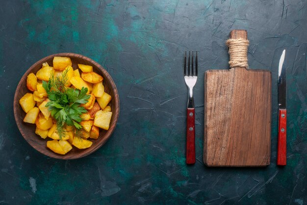 Vista superior de deliciosa comida de patatas en rodajas cocidas con verduras dentro de la placa marrón en el escritorio azul oscuro