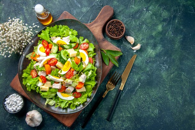 Vista superior de la deliciosa ensalada con ingredientes frescos en la tabla de cortar de madera, especias, botella de aceite, ajos, cubiertos, sobre fondo negro de colores de mezcla