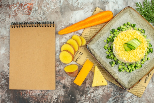 Vista superior de una deliciosa ensalada en un periódico viejo y dos tipos de queso y zanahorias, papas picadas, cuaderno en mesa de colores mezclados