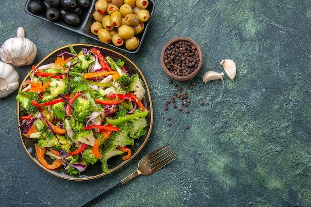 Vista superior de una deliciosa ensalada vegana en un plato con varias verduras y un tenedor, pimienta, aceitunas negras verdes, ajo en el lado derecho sobre fondo oscuro