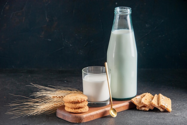 Vista superior de la deliciosa leche en vaso y botella, cuchara dorada, galletas apiladas en picos de bandeja de madera sobre superficie oscura