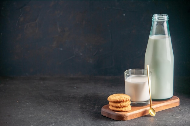 Vista superior de deliciosa leche en vidrio y botella cuchara dorada galletas apiladas en bandeja de madera en el lado izquierdo sobre fondo oscuro