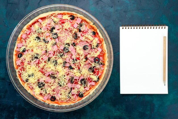 Vista superior deliciosa pizza de queso con aceitunas, salsa de tomate, salchichas dentro de una bandeja de vidrio con bloc de notas en el escritorio azul claro.