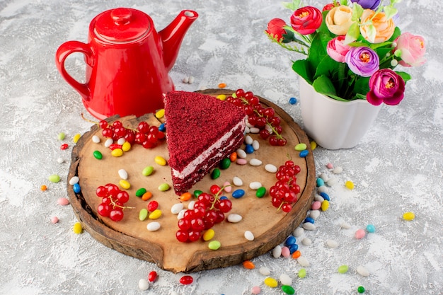 Vista superior deliciosa rebanada de pastel con crema y frutas junto con hervidor de agua rojo y flores en el escritorio de madera con coloridos caramelos pastel galleta té dulce
