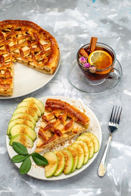 Una vista superior de la deliciosa rebanada de pastel de manzana dentro de la placa con té en el escritorio gris pastel de azúcar