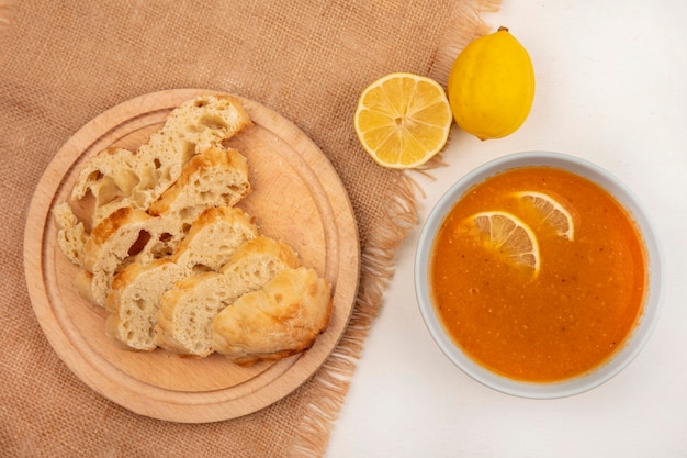 Vista superior de la deliciosa sopa de lentejas en un cuenco con rebanadas de pan sobre una tabla de cocina de madera sobre una tela de saco con limones sobre una superficie blanca