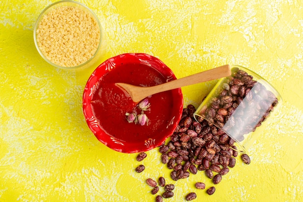 Vista superior de la deliciosa sopa de tomate dentro de la placa roja con frijoles crudos en la mesa amarilla, comida de sopa cena comida vegetal