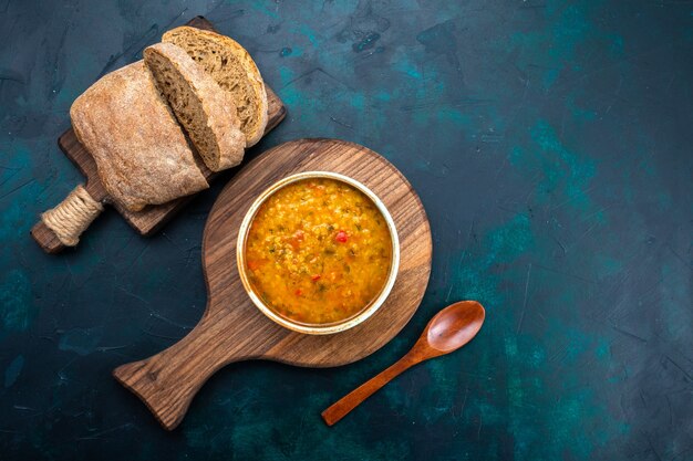 Vista superior deliciosa sopa de verduras dentro de un plato redondo con pan sobre el escritorio azul oscuro.