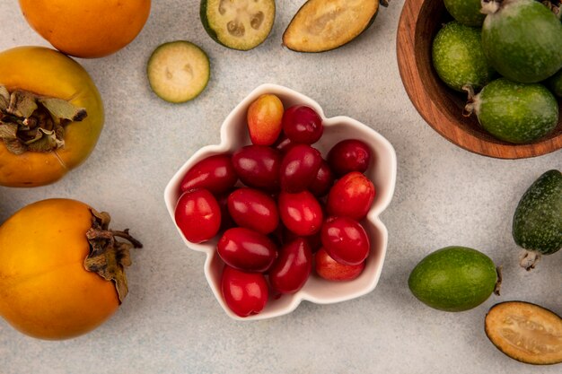 Vista superior de deliciosas cerezas de cornalina en un recipiente con feijoas y caquis aislado sobre un fondo gris