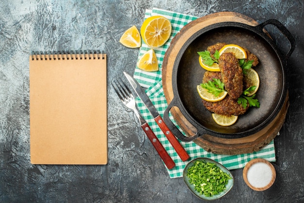 Vista superior de deliciosas chuletas servidas con verduras y limón en una sartén negra sobre cubiertos de madera sobre una toalla verde pelada sobre fondo de color oscuro con espacio libre