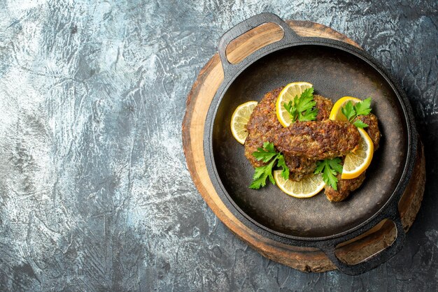 Vista superior de deliciosas chuletas servidas con verduras y limón en una sartén negra sobre una tabla de madera en el lado izquierdo sobre fondo de color oscuro con espacio libre