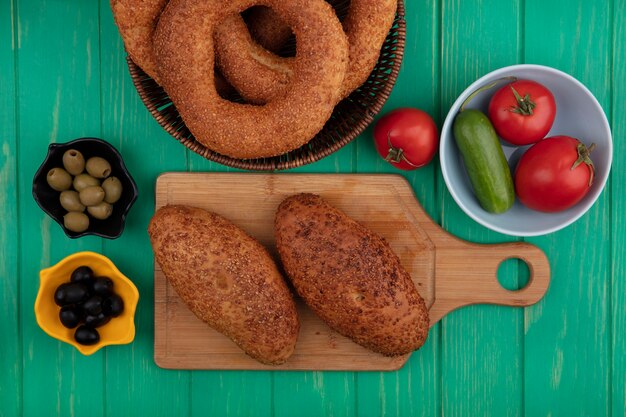 Vista superior de deliciosas y empanadas de sésamo en una tabla de cocina de madera con aceitunas en un recipiente con verduras en un recipiente sobre un fondo de madera verde