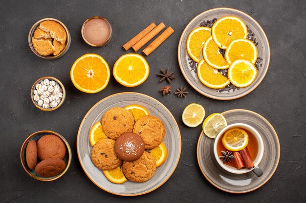 Vista superior deliciosas galletas de arena con naranjas frescas y una taza de té sobre fondo oscuro galleta de fruta galleta dulce azúcar cítrico