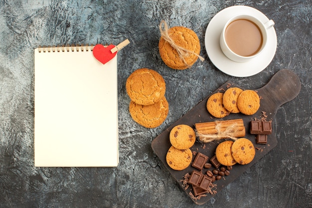 Vista superior de deliciosas galletas, barras de chocolate y una taza de café, cuaderno de espiral sobre una superficie oscura helada