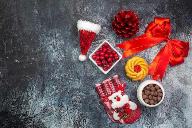 Vista superior de deliciosas galletas y chocolate cornel en ollas blancas calcetín de año nuevo cono de conífera roja cinta roja en el lado izquierdo sobre la superficie oscura