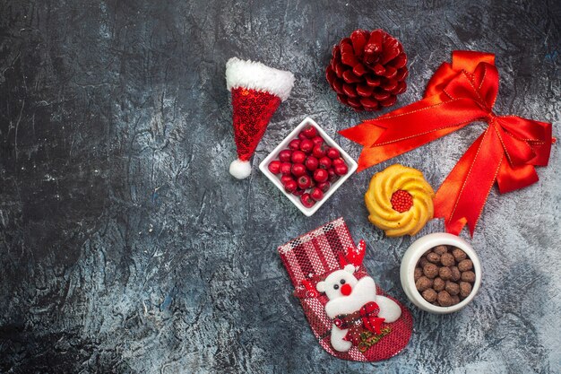 Vista superior de deliciosas galletas y chocolate cornel en ollas blancas calcetín de año nuevo cono de conífera roja cinta roja en el lado izquierdo sobre la superficie oscura