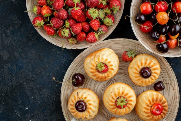 Foto gratuita una vista superior deliciosas galletas con fresas rojas cerezas dentro de la placa en el escritorio oscuro galleta cookie