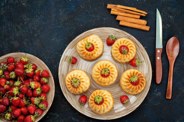 Una vista superior deliciosas galletas con fresas rojas dentro de la placa blanca en el escritorio oscuro hornear galletas