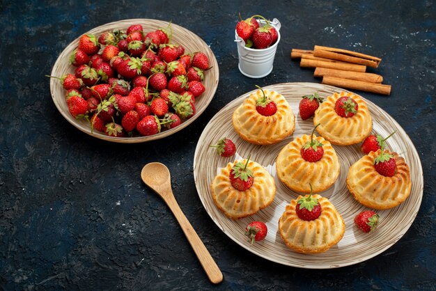 Una vista superior deliciosas galletas con fresas rojas dentro de la placa blanca en el té de galletas de escritorio oscuro