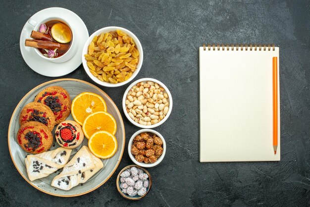 Vista superior de deliciosas galletas con pasteles afrutados, té de naranja y nueces en la superficie oscura, té de bocadillos, dulces dulces