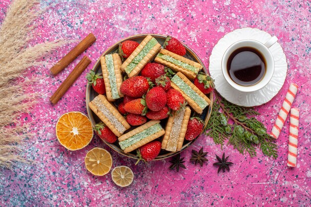 Vista superior de deliciosas galletas waffle con fresas rojas frescas y taza de té en superficie rosa